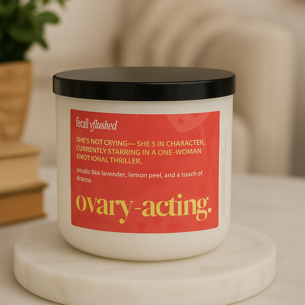 Candle with a red label on a marble surface, with a plant and books in the background.