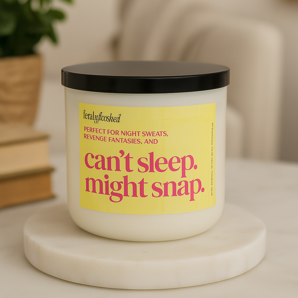 Candle with a humorous label on a marble coaster, surrounded by books and a plant.